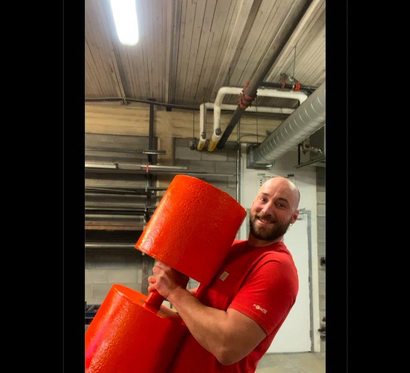 A person holding a large bright-orange circus dumbbell inside a gym workshop, showcasing heavy-duty strongman equipment used for strength and CrossFit training.